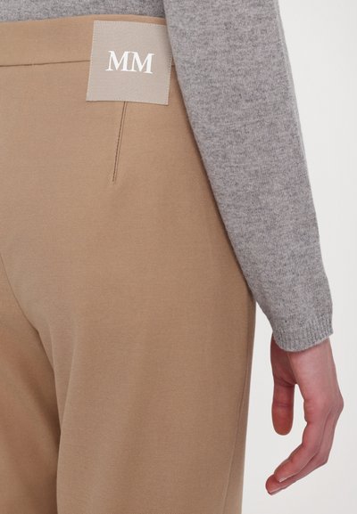 Beige Hosen mit glatter Textur und einer sichtbaren Gesäßtasche. Ein grauer Pulloverärmel ist sichtbar, mit einem hellgrauen Etikett, auf dem "MM" steht.