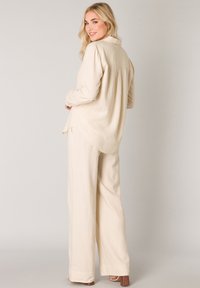 Beige, locker sitzendes Leinenhemd mit Kragen, langen Ärmeln und geschwungenem Saum, kombiniert mit passenden weiten Hosen. Strukturierter Stoff.