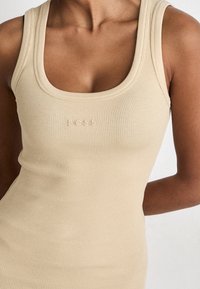 Kvinna iklädd en beige ribbad linnetopp med en rund halsringning och ordet "BOSS" broderat på bröstet.