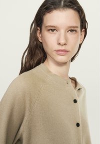 Cardigan beige realizzato in materiale morbido, con colletto rotondo, bottoni neri e maniche a raglan. Texture liscia con dettagli minimi.