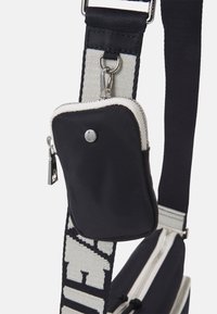 Borsa a spalla in tessuto nero con tasca con zip, dotata di una tracolla spessa con testo bianco a contrasto e dettagli in metallo argentato.