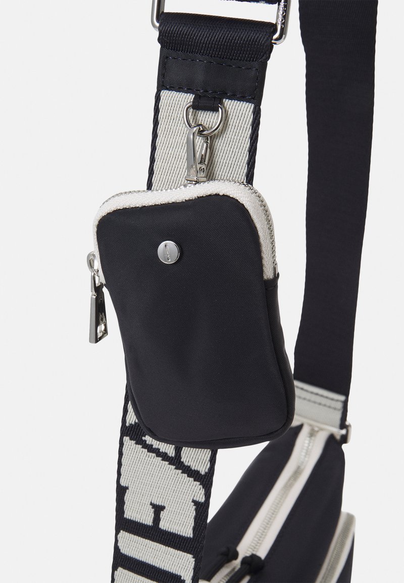 Schultertasche aus schwarzem Stoff mit einem Reißverschlussfach, ausgestattet mit einem dicken Trageriemen mit kontrastierendem weißen Text und silbernen Hardware-Details.