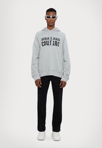 Γκρίζα hoodie με μαύρο κείμενο "VERSACE JEANS COUTURE", συνδυασμένο με μαύρο παντελόνι και λευκά αθλητικά παπούτσια. Μοντέλο που φοράει λευκά γυαλιά ηλίου.