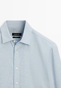 Camicia azzurra chiaro a bottoni realizzata in tessuto strutturato, con colletto classico e cinque bottoni bianchi sul davanti. Etichetta del marchio visibile.