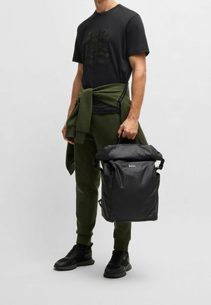 Mochila negra de tipo roll-top con correas ajustables, acabado mate, logo en la parte frontal, combinada con pantalones verdes oscuros y zapatillas negras.