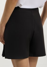 Parte superiore del corpo di una donna che indossa pantaloni corti neri a vita alta e una camicia bianca infilata, su uno sfondo semplice.