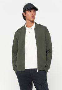 Olivengrøn zip-up jakke med rund hals, ribbede manchetter og sidelommer, båret over en hvid polo skjorte og sammen med mørke bukser. Sort kasket.