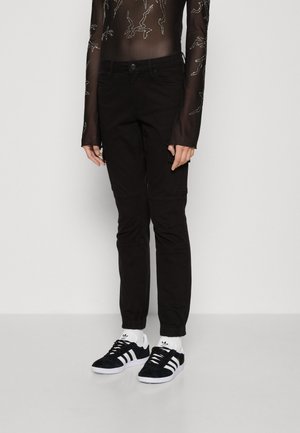 Personne portant un pantalon noir ajusté, des baskets Adidas noires et blanches, des chaussettes blanches montantes et un haut noir transparent à manches longues avec des motifs argentés.