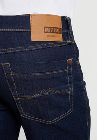 Tumed sinised teksad pruuni nahkpleki ja sildiga "MUSTANG True Denim", oranž õmblus ja eristuv tasku disain.