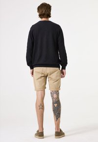 Zwarte trui, lichte kaki shorts, zichtbare tatoeage op het linker been, en bruine schoenen. De figuur is van achteren afgebeeld tegen een effen achtergrond.