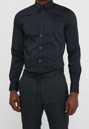 Uomo che indossa una camicia nera aderente con bottoni infilata in pantaloni a quadri grigio scuro con coulisse, su uno sfondo semplice.