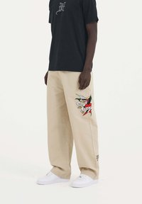 Beżowe spodnie szerokie z haftowanym motywem przedstawiającym ptaka oraz banerem z napisem "PRAWDZIWA ŚMIERĆ". Połączone z czarną koszulką i białymi trampkami.