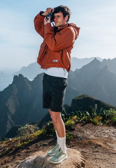 Person klädd i en orange huva-jacka, svarta shorts och ljusgröna sneakers, som står på stenigt terräng med berg i bakgrunden.