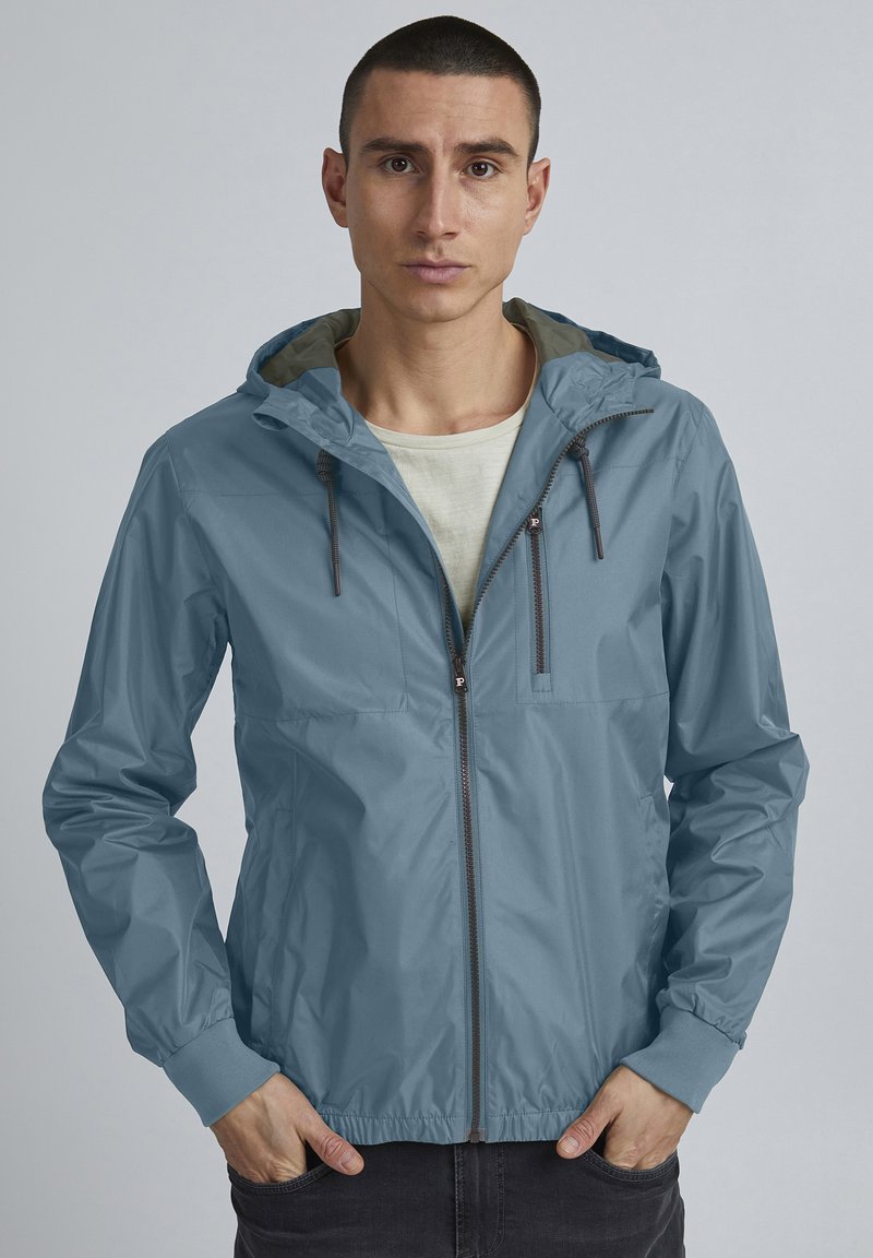 Veste imperméable bleu clair avec une capuche à cordon, fermeture éclair à l'avant et poignets élastiques, portée par-dessus une chemise de couleur claire.