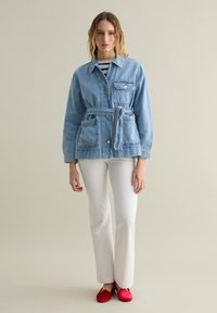 Giacca di jeans azzurro chiaro con vita aderente, tasca applicata, chiusura con bottoni e maniche lunghe, abbinata a pantaloni bianchi e scarpe rosse.