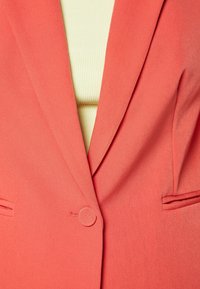 Koralblazer med skräddarsydd design, med slagkragar och en enkel knappstängning. Tyget verkar slätt, kompletterat av en ljusgul topp under.