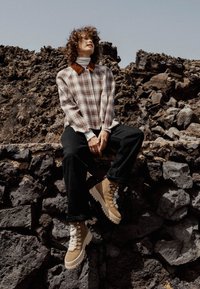 Una camicia a quadri marrone con colletto marrone, pantaloni neri e stivali beige con lacci posata su un muro di roccia contro un paesaggio vulcanico accidentato.