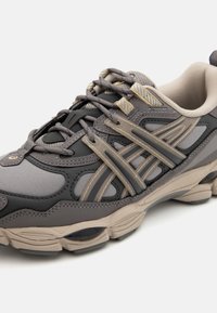 Chaussure de sport grise et beige comprenant des matériaux en mesh et synthétiques, des bandes contrastantes, un bout rond et une semelle rembourrée avec des détails de traction.