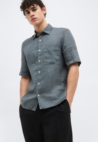 Νέος άνδρας που φοράει γκρι πουκάμισο με κοντά μανίκια και τσέπη στο στήθος, καθώς και μαύρο παντελόνι, στέκεται με τα χέρια στις τσέπες.