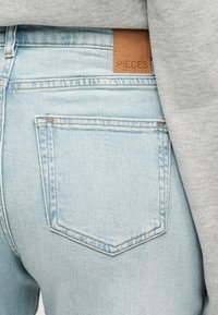 Πίσω τσέπη ανοιχτό μπλε τζιν με ταμπέλα από δέρμα σε χρώμα ταμπά "PIECES" στη μέση, σε συνδυασμό με γκρι τοπ.