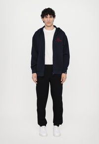 Marinblå zip-hoodie med röd text, tillsammans med en vit t-shirt och svarta sweatpants. Modellen bär vita sneakers; avslappnad design och passform.