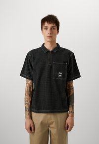 Must have lühikeste varrukatega polo särk, millel on valged vertikaalsed triibud, kaelus, poolne tõmblukk ja eesmine tasku, millel on logo kleebis.