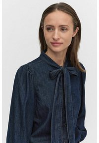 Mörk denimblus med en knytbåge detaljerad vid halsen, långa ärmar med samlade manschetter och knappstängning fram. Mjuk textur och klassisk design.