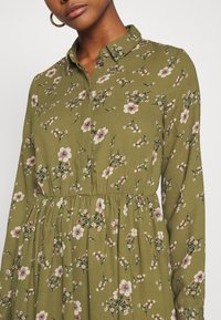 Olijfgroene bloemenjurk met lange mouwen, een knoopsluiting bij de kraag en een gathered taille. Heeft een patroon van roze en witte bloemen op een textuurstof.