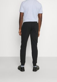 Schwarze Jogginghosen mit schmalem Schnitt und elastischem Bund, kombiniert mit einem hellblauen Hemd und schwarzen Schuhen. Glattes Material, minimalistische Details.