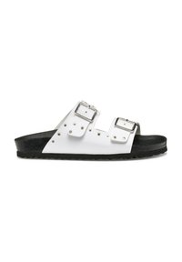 Sandalias blancas de dos correas con hebillas plateadas, suela texturizada negra y tachuelas decorativas a lo largo de las correas. Diseño plano con punta abierta.