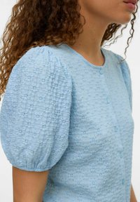 Blusa textured blu chiaro con maniche a sbuffo e scollo rotondo, dotata di piccoli bottoni e un motivo sottile su tutta la stoffa.