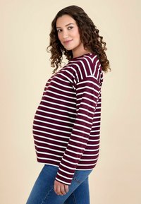 Tricou de maternitate cu mâneci lungi, în dungi maronii și albe, cu o croială lejeră și guler rotund, asortat cu blugi albaștri, pe un fundal neutru.
