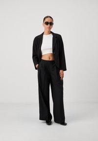 Black tailored blazer with a single button, paired with high-waisted black wide-leg trousers. Model wears a white crop top and dark sunglasses.