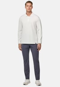 Polo shirt bianco a maniche lunghe con colletto, abbinato a pantaloni sportivi grigi e sneakers bianche. Texture liscia, vestibilità casual, dettagli minimi.
