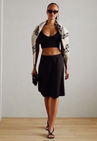 Sort crop top, sort knælang nederdel og sorte sandaler. Draperet sweater i en lys farve med et mønster. Holder en lille taske.