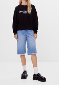 Svart sweatshirt med vit tryck, ljusblå denimshorts med fransad kant, och svarta klumpiga skor i kombination med vita strumpor.