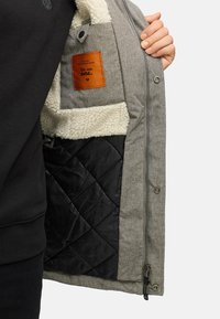 Gray outer jacket with faux shearling lining and black quilted inner pocket. Features orange label with text. Zipper and snap button closure.