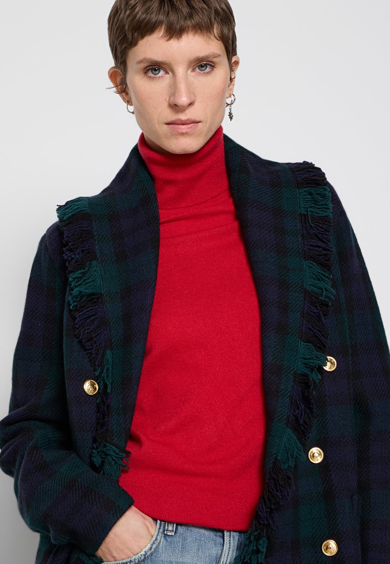 Marynarka w kratę z wełny w ciemnozielonym i granatowym kolorze, z złotymi guzikami i frędzlami na krawędziach. Nosi ją z czerwoną golfem.