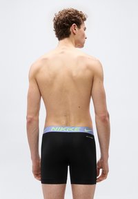 Άνδρας μοντέλο, φωτογραφημένος από πίσω, φοράει μαύρα ανδρικά μποξεράκια Nike με μπλε ζωνάρι στη μέση και λογότυπο νέον πράσινο, σε απλό λευκό φόντο.