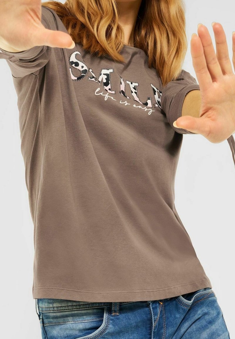 Brun långärmad skjorta med en grafisk "SMILE"-design i djurmotiv. Mjuk tygkvalitet, rund halslinning, kombinerad med blå jeans.