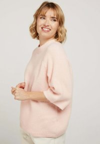 Pull rose clair texturé à manches courtes et à encolure ronde, porté avec un pantalon de couleur neutre. Matière douce et pelucheuse.