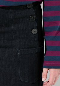 Tmavě modrá džínová sukně se rovnými liniemi, s třemi černými knoflíky po boku a náprsní kapsou. Viditelný pruhovaný top s dlouhým rukávem.