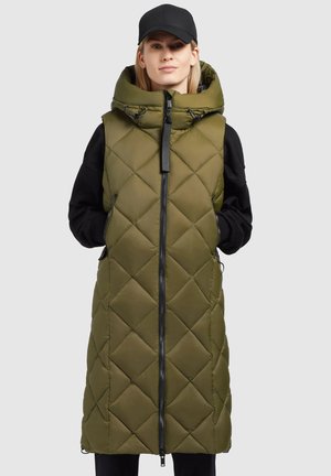Femme portant un long gilet matelassé sans manches vert olive avec un grand col et une casquette noire, les mains dans les poches, se tenant devant un fond uni.