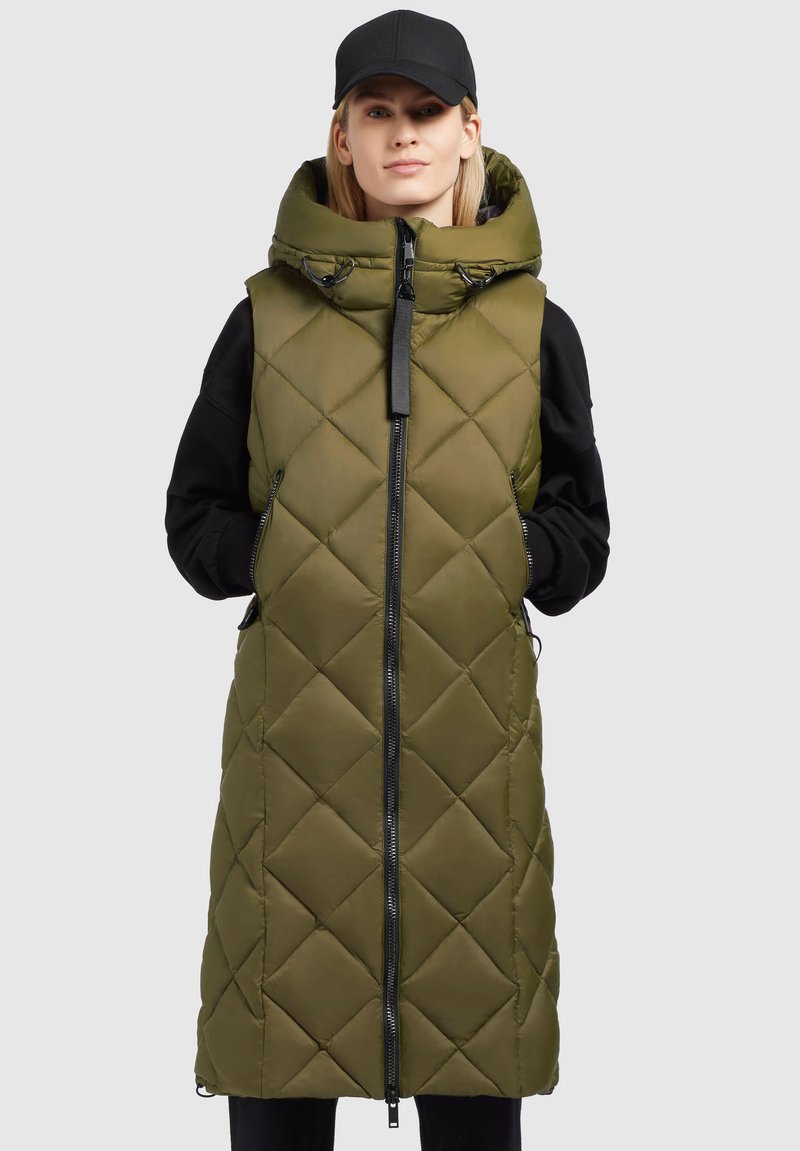 Frau, die eine langärmlige, olivgrüne, gesteppte Weste ohne Ärmel mit großem Kragen und schwarzem Hut trägt, mit Händen in den Taschen, steht vor einem schlichten Hintergrund.