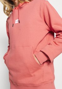 Růžová mikina s kapucí vyrobená z měkké tkaniny, s přední kapsou, žebrovanými manžetami a malou značkovou nášivkou poblíž výstřihu.
