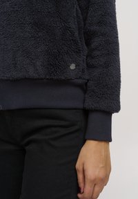 Mørkeblå fleece-genser med teksturert overflate og ribbede mansjetter. Har en liten sirkulær logo nede til venstre nær sømmen.