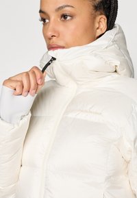 Biela izolovaná pufrová bunda s vysokým golierom, zipsom na uzatvorenie a detailom loga. Vyznačuje sa mäkkou tkaninou a štíhlym dizajnom.