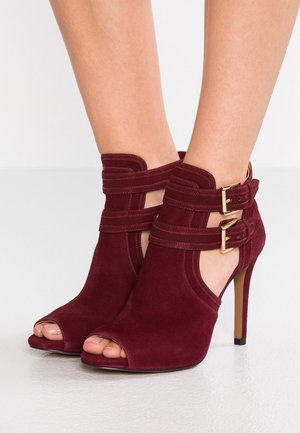 Bottines à talons hauts en daim bordeaux avec orteils ouverts et doubles brides à boucle dorée, portées pieds nus sur un fond blanc.