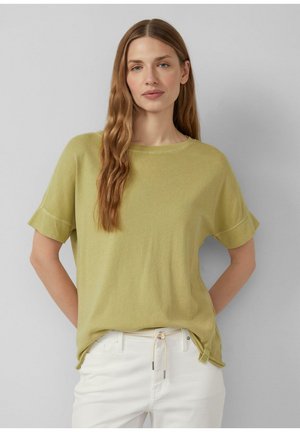 Vrouw met lang golvend haar, gekleed in een lichtgroen topje met korte mouwen en een witte broek met trekkoord, tegen een effen grijze achtergrond.