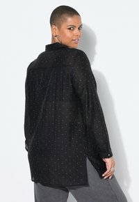 Schwarze, durchsichtige Bluse mit einem gefaltetem Design, verziert mit kleinen weißen Punkten. Mit Kragen und langen Ärmeln, sowie seitlichen Schlitzen am Saum.